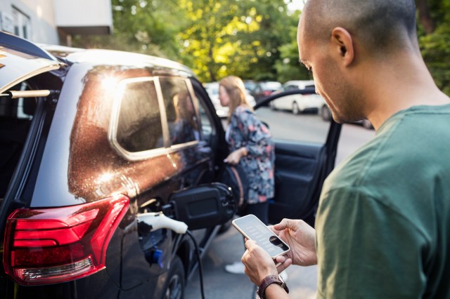 Man using smartphone with woman standing by car in background