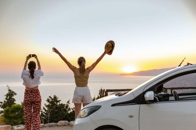 Rear view of two young women taking a break during a road trip. They are standing on a viewpoint and looking at the sunset
